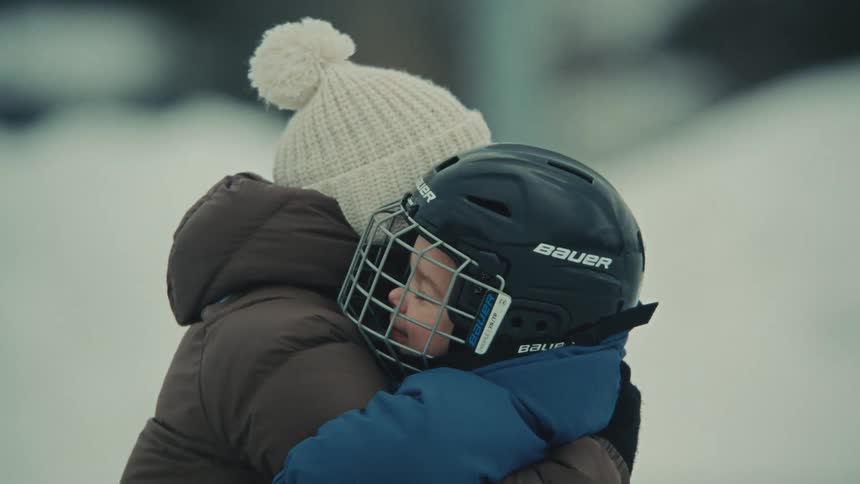 A mother warmly hugs her son wearing a hockey helmet on a snowy day, sharing a tender moment
