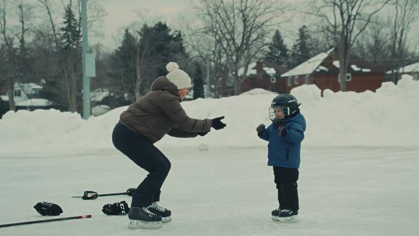 A patient mother encourages her young son as he learns to ice skate on a snowy outdoor rink