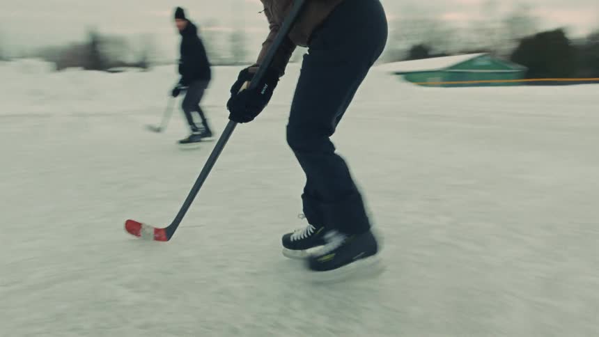 A dynamic slow-motion shot follows a person ice skating and handling a puck while playing hockey with a friend on an outdoor rink