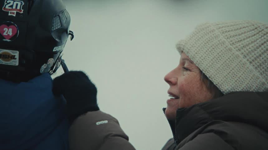 A smiling woman in a knit hat lovingly adjusts a child's hockey helmet on a snowy day