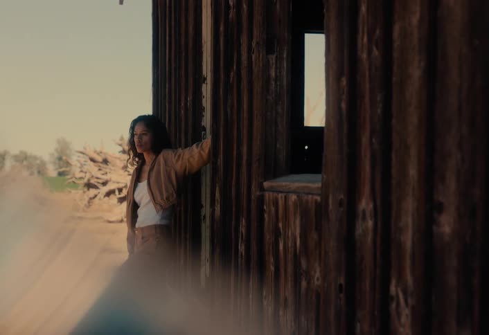 A pensive young woman emerges from a rustic wooden shack and walks away in the warm glow of sunset