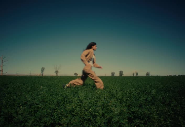 A woman with long dark hair runs in slow motion from left to right across a wide, lush green field under a clear blue sky