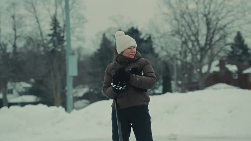 A smiling older woman puts on her gloves to play hockey with a younger man on an outdoor rink in the snow