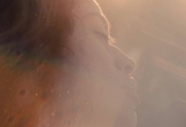 In a hazy, sun-drenched slow-motion close-up, a woman's face in profile appears contemplative and hopeful
