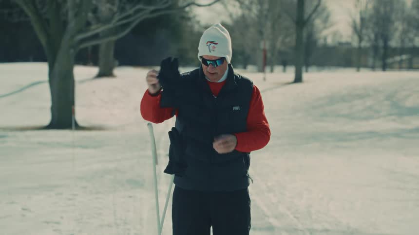 An elderly man prepares for a day of cross-country skiing, putting on his hat, sunglasses, and gloves in a sunlit, snowy park