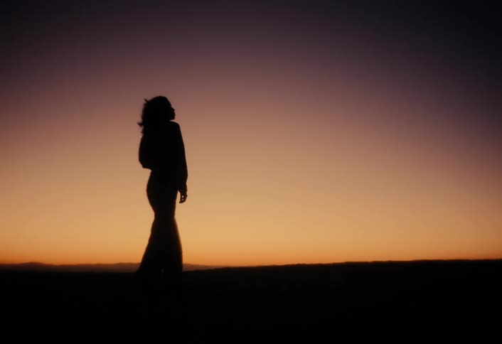 A woman's silhouette walks in slow motion across a hilltop against the beautiful gradient of a sunset sky