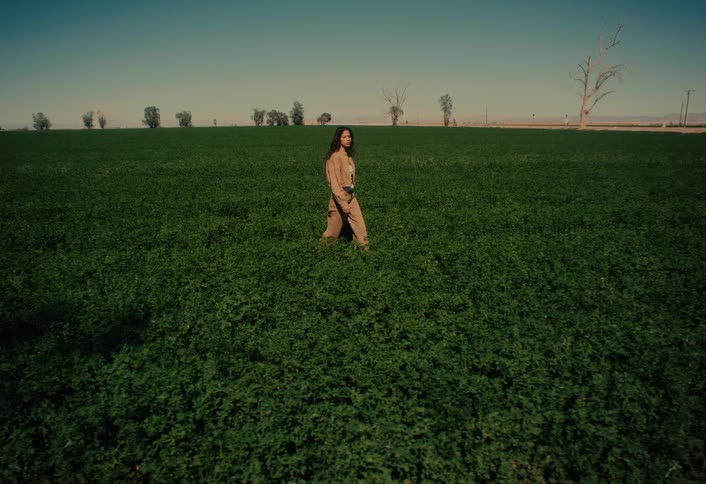 Cinematic slow motion shot of a woman in a tan jumpsuit walking through a lush green field on a bright, sunny day