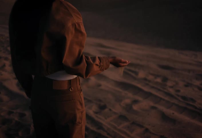 In slow motion, a woman standing alone on a vast desert sand dune at sunset lets the fine sand sift through her fingers