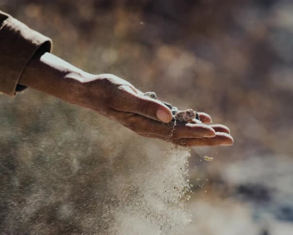 A close-up shot of an open hand letting dirt and rocks fall through its fingers in slow motion as sunlight flares