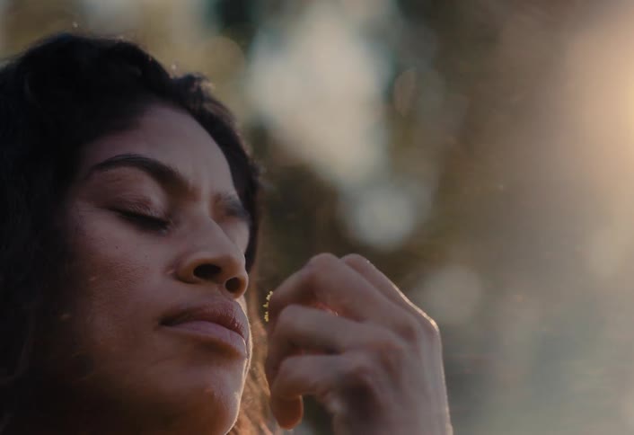 A serene woman with her eyes closed enjoys the gentle scent of a wildflower in slow motion as the golden hour sun flares behind her