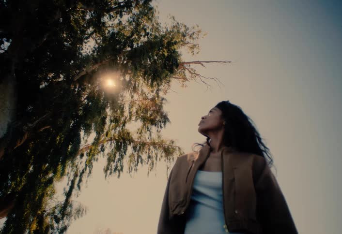 A thoughtful woman in a brown jacket gazes up at a large tree as the bright sun shines through its leaves, creating a beautiful flare