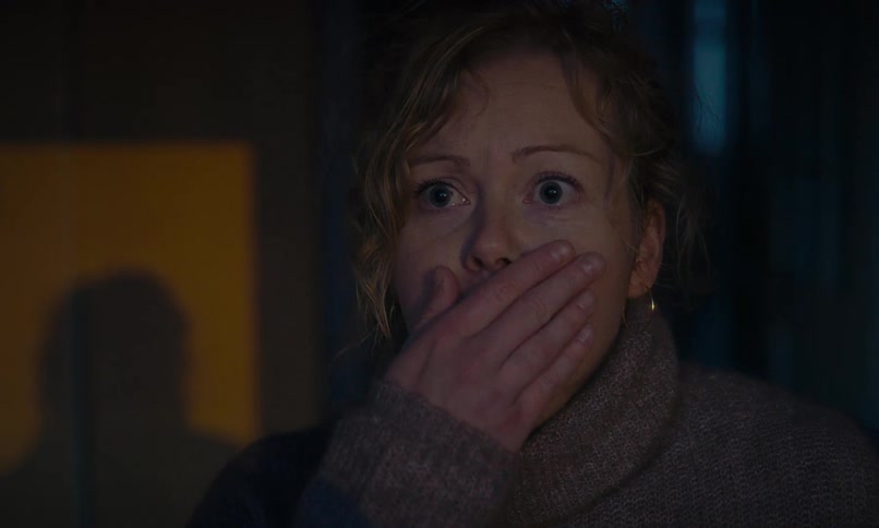 A terrified woman peeks out from the shadows in a dark room and covers her mouth in absolute shock and horror