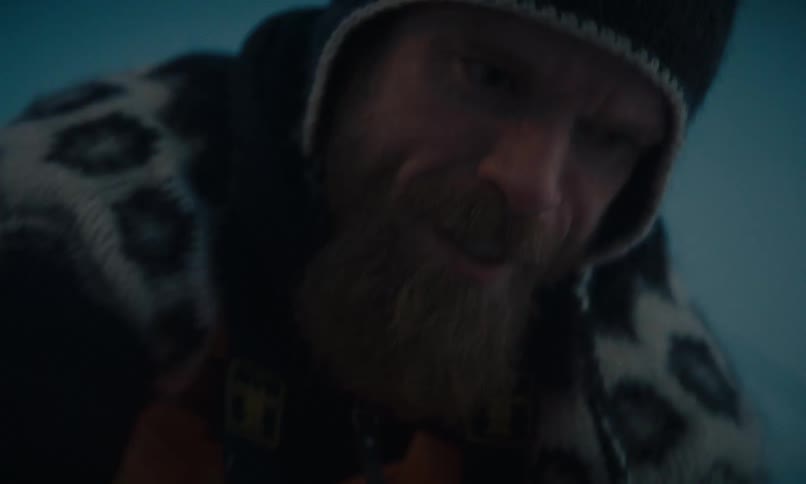 A close-up shot of a bearded man in a beanie and warm clothing concentrating on a task in a dim, cold environment