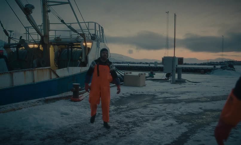 A fisherman in bright orange overalls gets off his boat and walks across a snowy pier to talk with a colleague at dusk