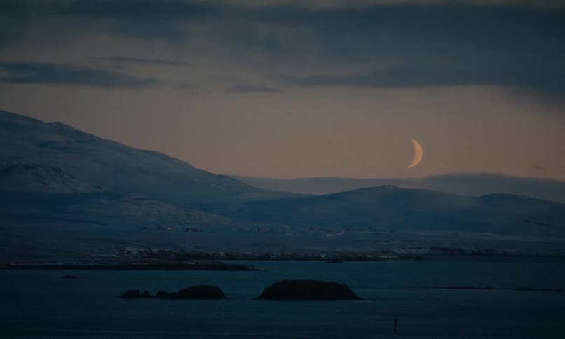 A beautiful crescent moon hangs in the twilight sky over a tranquil bay and snow-covered mountains