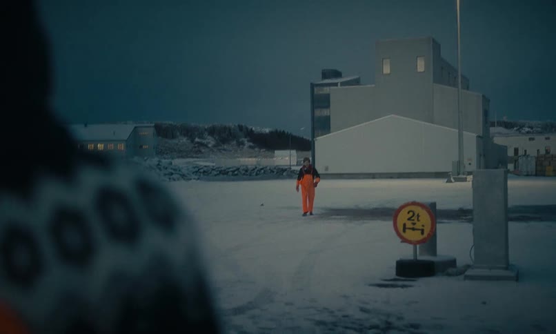 A Norwegian fisherman in bright orange overalls walks across a snowy port at dusk towards a coworker