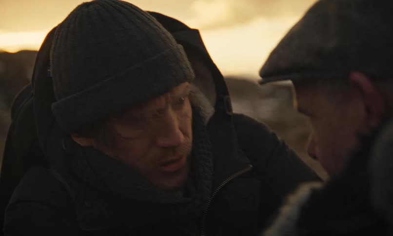 A desperate and injured man argues intensely with another man as they struggle together in a cold, desolate landscape at dusk