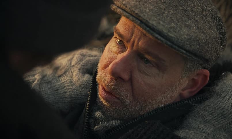 A tense close-up shows a man in a flat cap and winter clothes looking up with fear as he speaks to someone off-camera