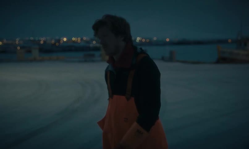 A weary fisherman in orange overalls walks in slow motion across a snow-covered dock at night