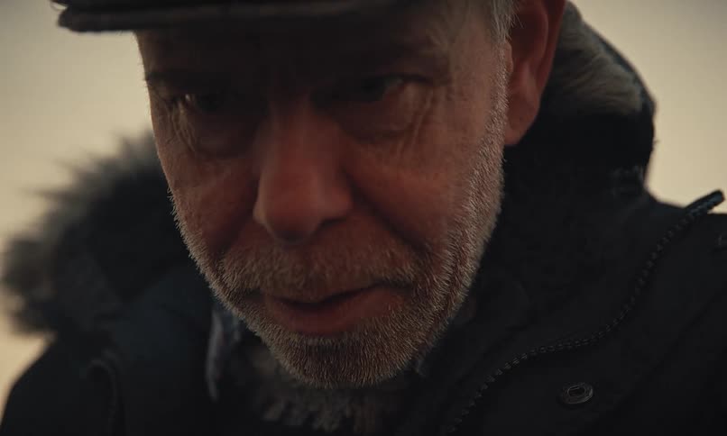 A dramatic close-up reveals the focused face of an elderly man wearing a cap and warm winter jacket