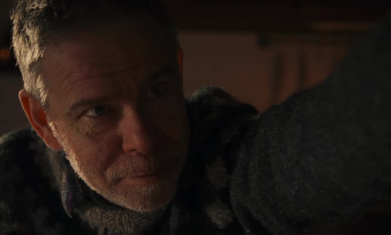 A close-up shot of a middle-aged man in a warm sweater, whose serious expression melts into a playful smile and a wink in a cozy, candlelit room