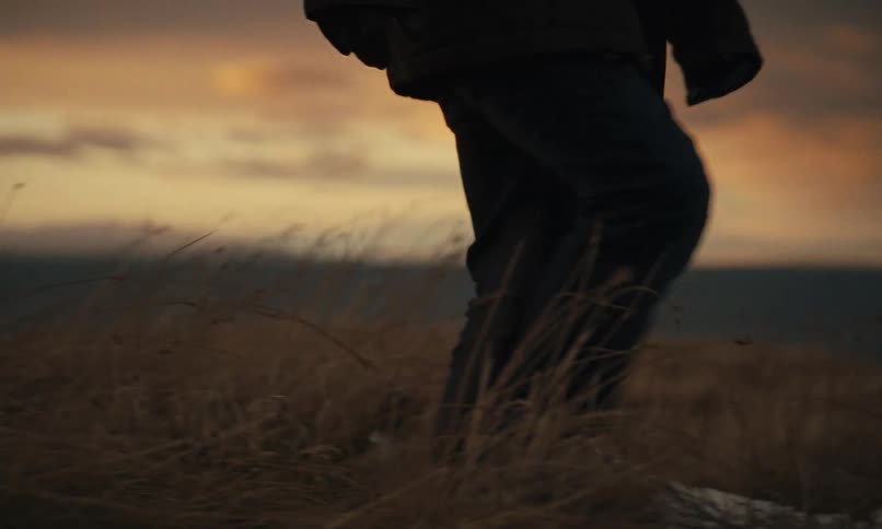 An exhausted man collapses onto the snowy ground of a desolate field overlooking the ocean at sunset