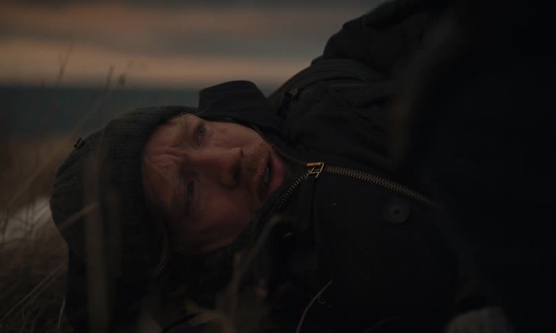 A close-up shot of a wounded and shivering man lying on the frozen ground while being comforted by another person