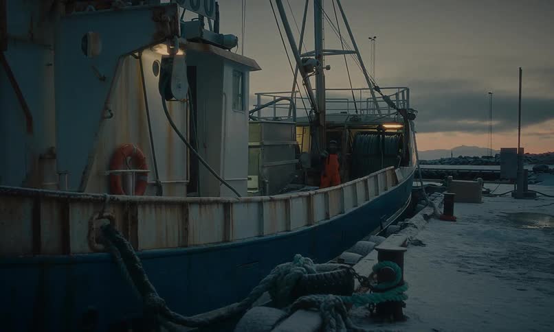 A lone fisherman works on the deck of a rusty boat docked in a snowy harbor at twilight