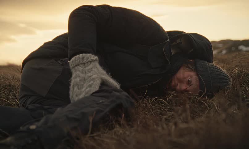 A dramatic close-up of an injured person wearing winter clothes, lying motionless in a desolate, grassy field at dusk