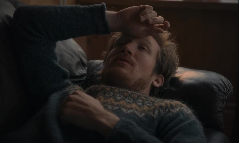 A handsome man in a patterned sweater lies back on a leather couch in a dimly lit room, appearing thoughtful and relaxed