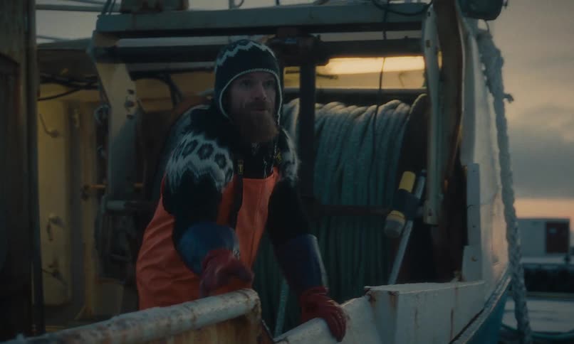 A rugged Icelandic fisherman in warm overalls walks along the frosty deck of his boat in a snowy harbor at dawn