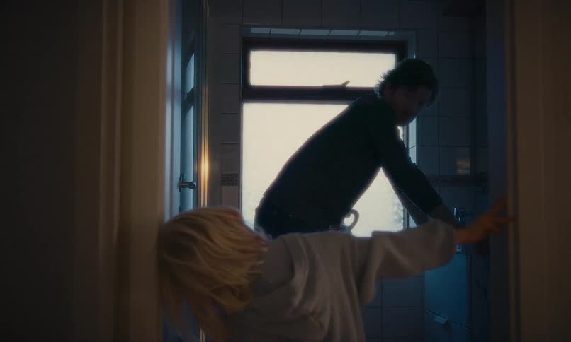 A worried woman in a robe watches a distressed man in a dark, backlit bathroom as he slowly turns to meet her gaze