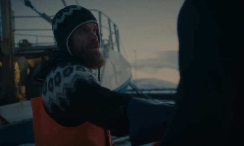Two rugged fishermen in warm winter gear work together on the snowy deck of their boat in a harbor at dusk