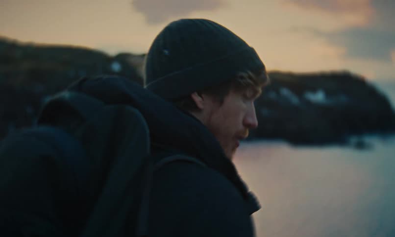 A solitary hiker in a beanie and winter coat gazes thoughtfully before embarking on a journey along a snowy shore at dusk