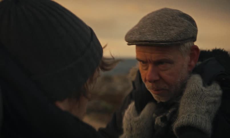 A senior man in a flat cap has a serious and heartfelt conversation with another person in a cold, windswept landscape at dusk