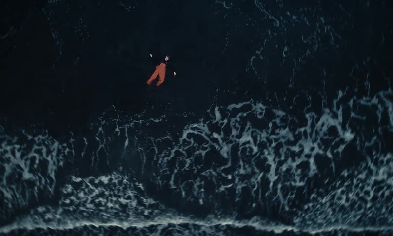 An aerial top-down view of a person in orange pants floating in the dark, foamy ocean waves of a black sand beach