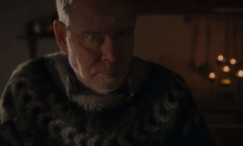 A serious older man with a pensive expression sits at a rustic table with two steaming mugs in a dimly lit, cozy room