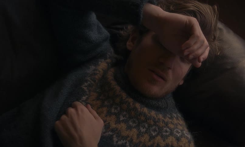 A weary young man in a wool sweater lies down with his arm over his eyes, appearing sick or depressed