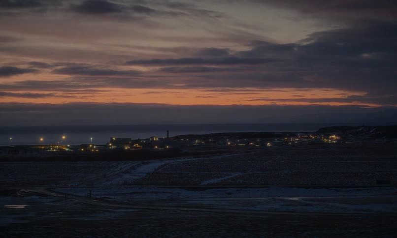 An illuminated coastal village sits on a snowy shoreline under a colorful sunset sky