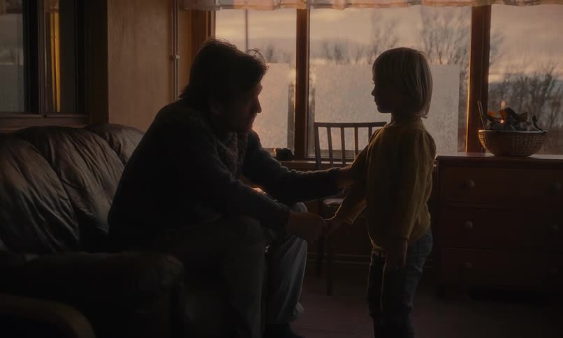 A young son holds his father's hand in a quiet, somber moment before walking away, leaving the man alone in a dimly lit room