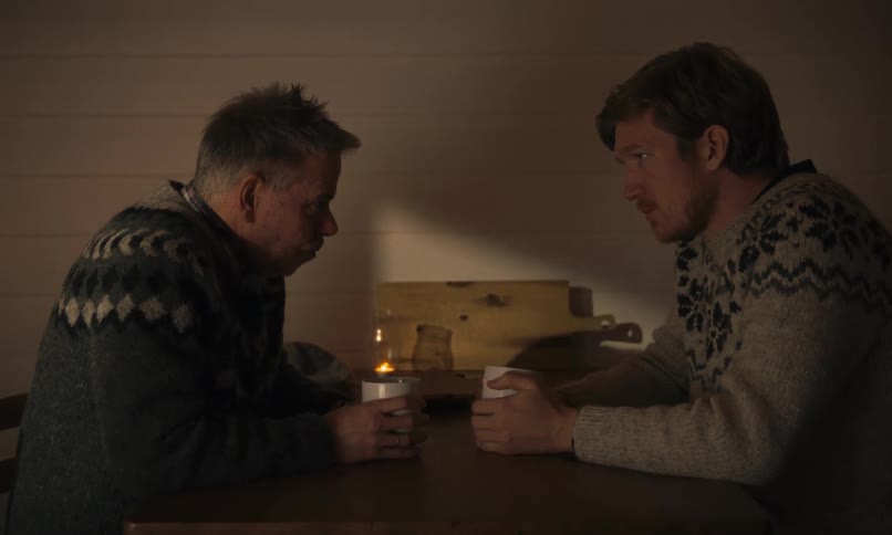 Two men in Nordic sweaters sit in thoughtful silence at a wooden table, drinking from mugs in the warm glow of candlelight