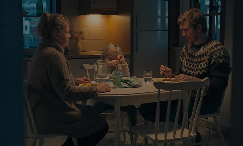 A young child cries at the dinner table while their concerned parents sit with them in a dimly lit kitchen
