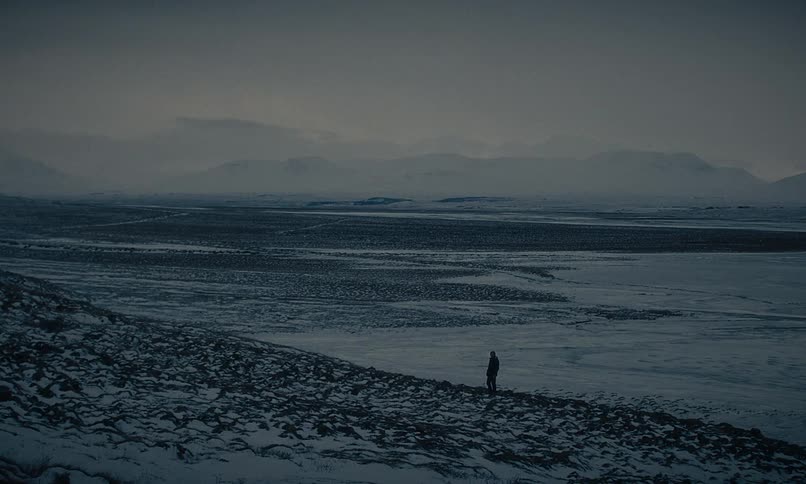 A solitary person walks along a snowy, rocky ridge overlooking a vast frozen valley at dusk