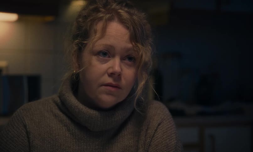 A tired and emotional woman with curly hair listens and speaks with a somber expression in a dimly lit kitchen