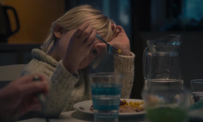 A tired young boy with blond hair plays with his food instead of eating during a dimly lit family dinner