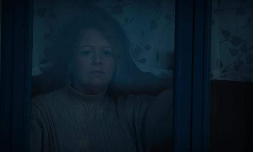 A lonely woman with a sad expression appears from the darkness to look out her window at night