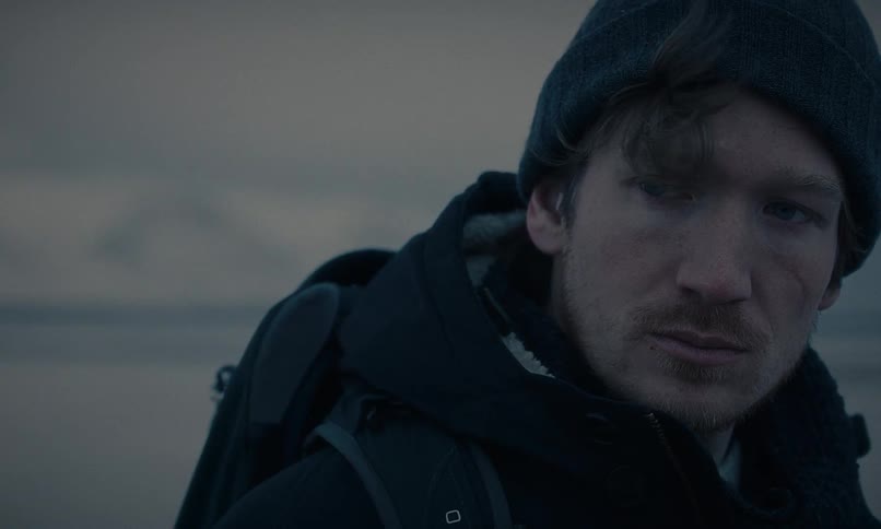Close-up of a rugged explorer in a winter coat and beanie turning to look intently at the camera in a bleak, cold environment