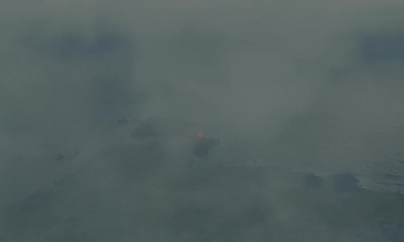 An atmospheric aerial view captures a lone lighthouse on a rugged peninsula as it is slowly consumed by encroaching fog at dusk