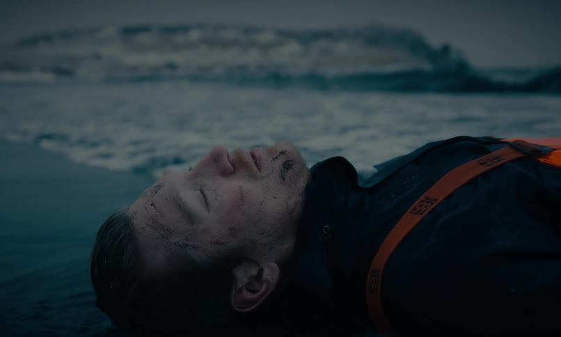 An unconscious man with a dirty face and wearing a life vest lies on a cold, desolate beach as waves wash over him
