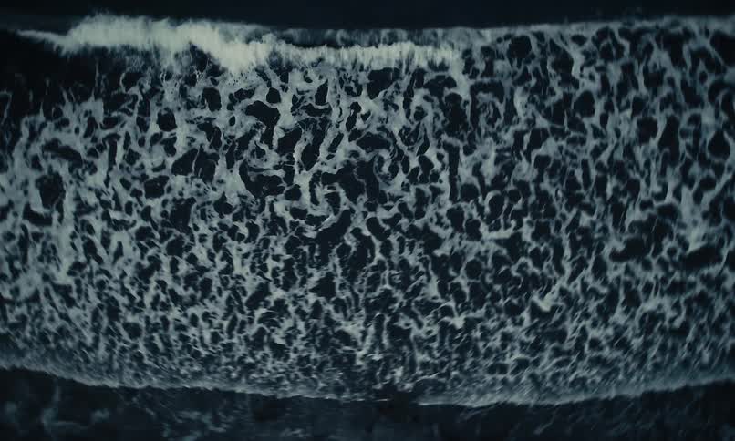 Hypnotic top-down view of dark ocean waves rolling onto a black sand shore in slow motion